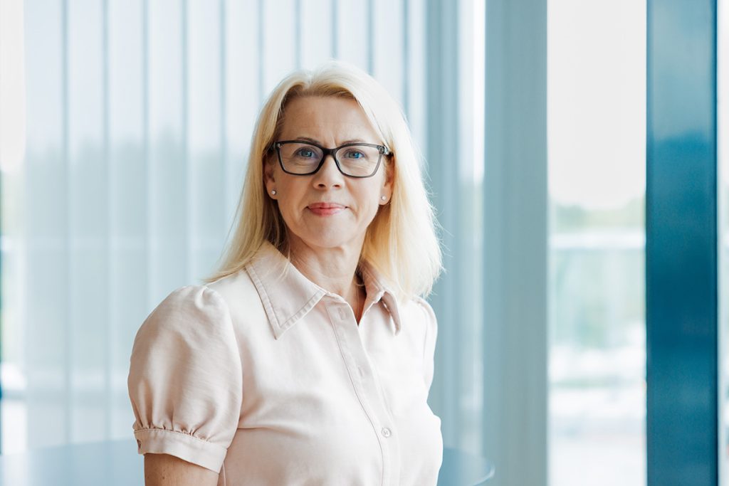 En middelaldrende kvinne med blondt hår og briller står innendørs, iført en lys rosa bluse. I bakgrunnen skimtes lamellgardiner og store vinduer.