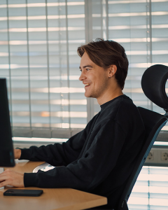 En person sitter ved et skrivebord og smiler mens han ser på en dataskjerm i et rom med persienner.