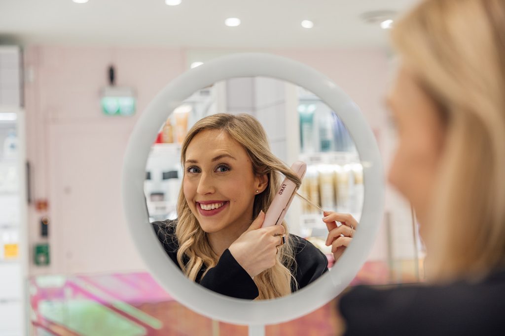 En kvinne styler håret med krølltang mens hun smiler til speilbildet sitt i et rundt speil i et sterkt opplyst rom. Alexandra er norgessjef i Lyko.