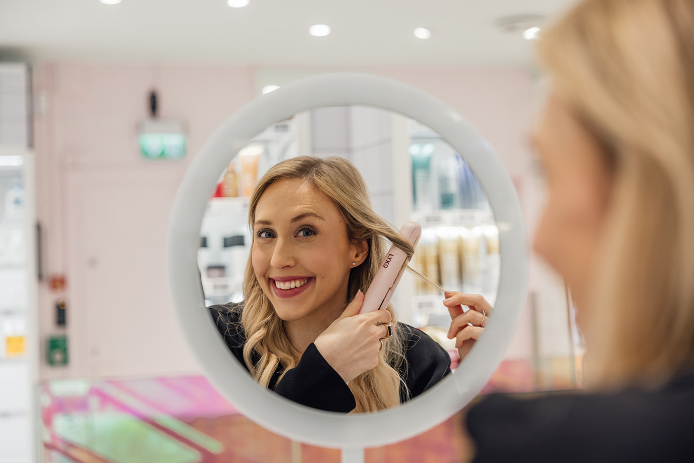 En kvinne styler håret med krølltang mens hun smiler til speilbildet sitt i et rundt speil i et sterkt opplyst rom. Alexandra er norgessjef i Lyko.