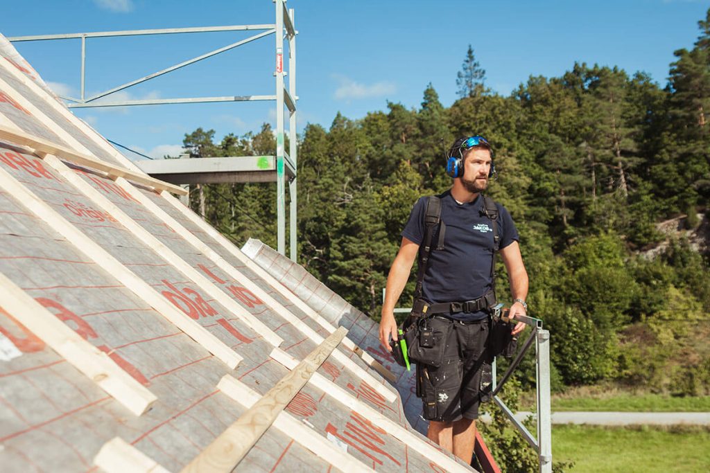 Kjell fra Byggmester Jakob er i gang med et nytt prosjekt. Han står på taket av et hus, med skog og blå himmel i bakgrunnen.