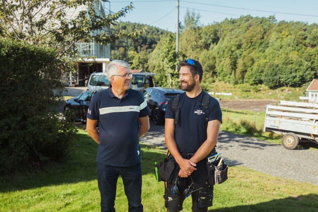 To menn står ute på en gressplen og snakker og smiler. I bakgrunnen ser vi trær, biler og et hus.