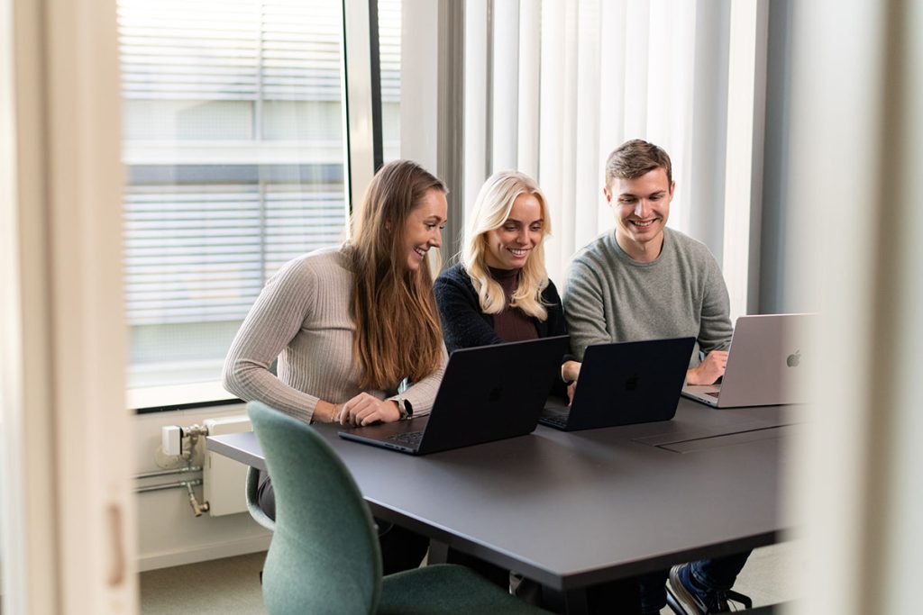 Tre personer sitter ved et bord med hver sin bærbare datamaskin og smiler. Illustrasjonsbilde til artikkel om partnermarkedsføring i Tripletex.
