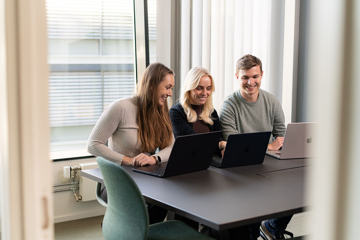 Tre personer sitter ved et bord med hver sin bærbare datamaskin og smiler. Illustrasjonsbilde til artikkel om partnermarkedsføring i Tripletex.