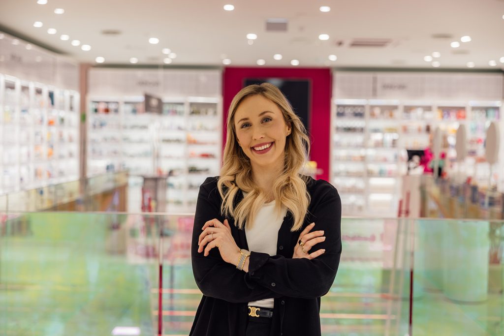 En kvinne med blondt hår står smilende med armene i kors i en sterkt opplyst butikk med varehyller i bakgrunnen.