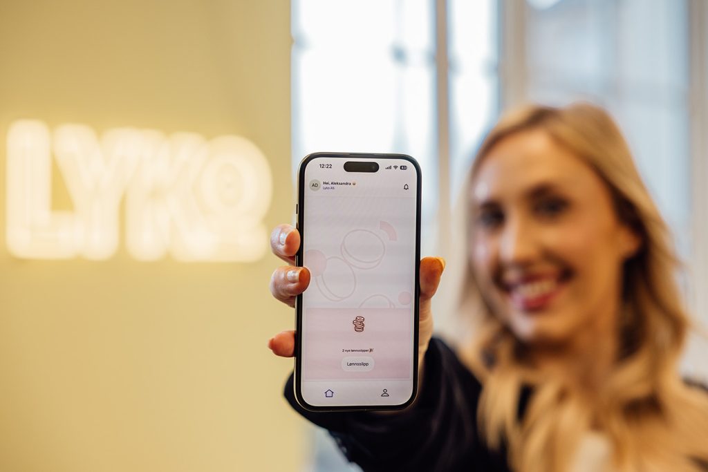 En person holder en smarttelefon som viser en appskjerm, med et uskarpt LYKO-skilt og vinduer i bakgrunnen.