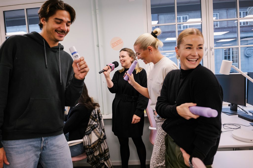 Fire personer på et kontor ler og synger inn i hårproduktflasker og en mikrofon, mens en annen person sitter ved en datamaskin i bakgrunnen.