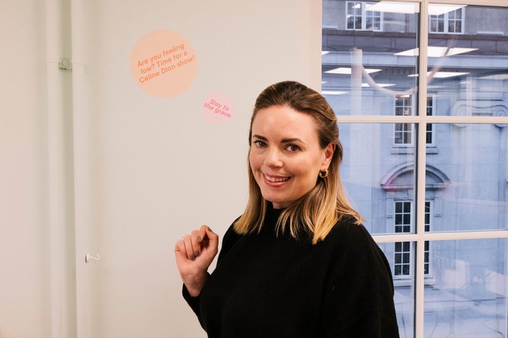 En kvinne med skulderlangt hår står innendørs ved et vindu og smiler til kameraet. På veggen bak henne står det: "Are you feeling low? På tide med et Celine Dion-show!.