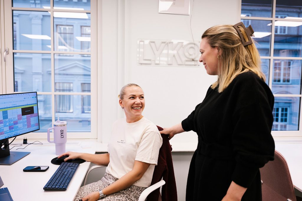 To kvinner samhandler på et moderne kontor; den ene sitter smilende ved et skrivebord med en datamaskin, mens den andre står ved siden av henne med en hånd på skulderen.