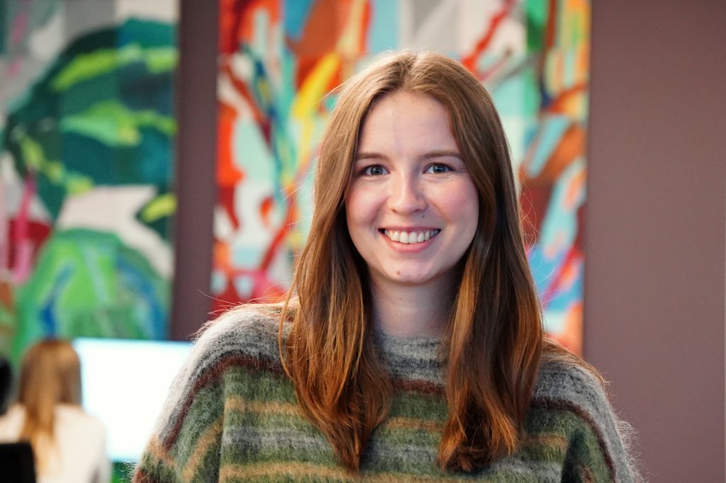 En ung kvinne med langt, brunt hår smiler til kameraet mens hun står innendørs foran et fargerikt, abstrakt maleri. Maren Aamodt leder teamet for storkundesupport i Tripletex.