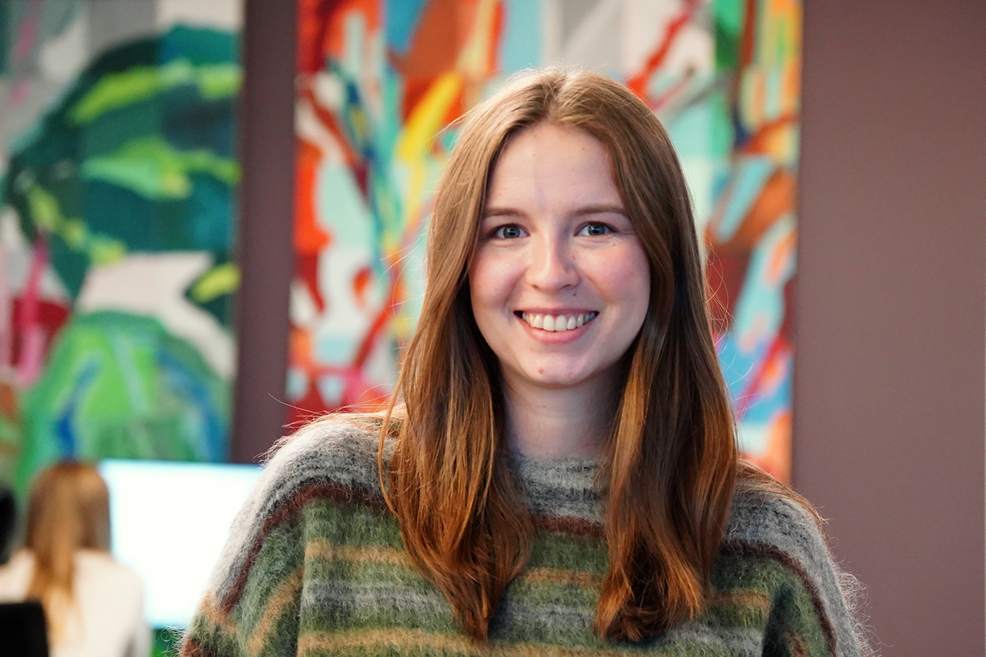 En ung kvinne med langt, brunt hår smiler til kameraet mens hun står innendørs foran et fargerikt, abstrakt maleri. Maren Aamodt leder teamet for storkundesupport i Tripletex.