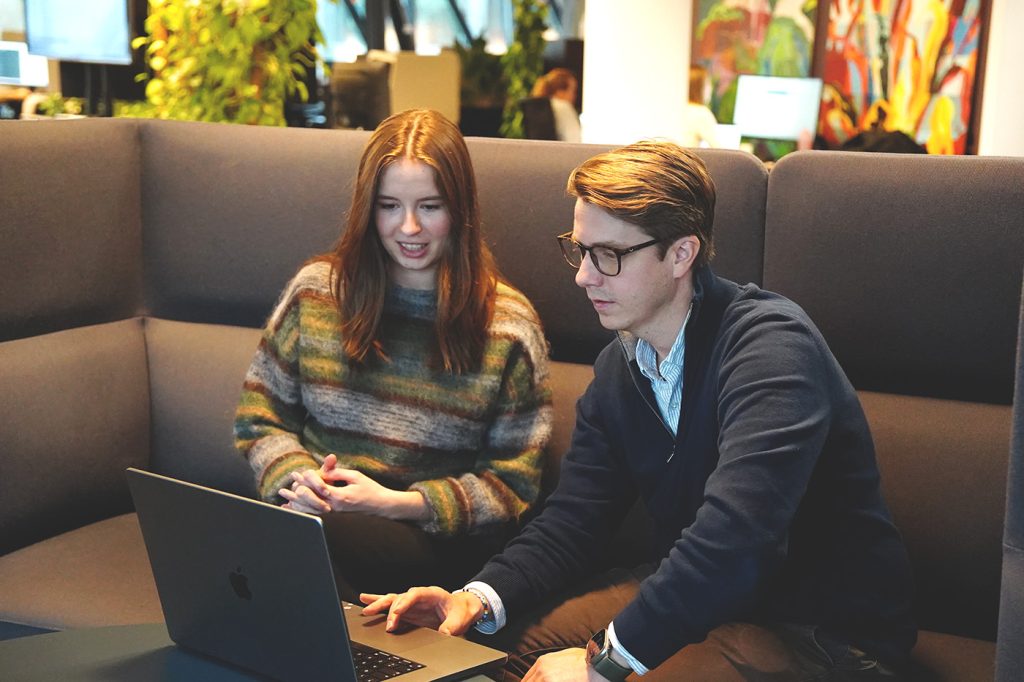 To personer sitter i en grå sofa og ser på en bærbar datamaskin sammen i et moderne kontor fylt med planter og fargerike kunstverk, og gir hverandre støtte mens de samarbeider.