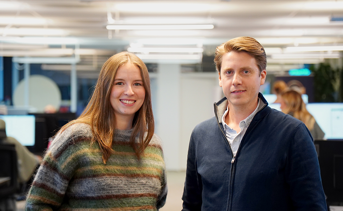 To personer står innendørs i et kontormiljø, vendt mot kameraet og smilende, med pulter og kolleger i bakgrunnen.