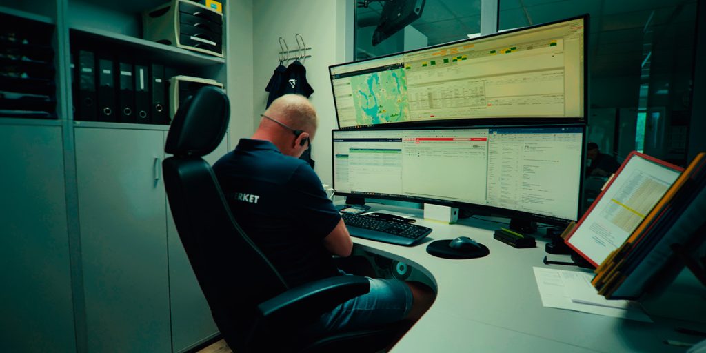 En person sitter ved et skrivebord på et kontor og arbeider med flere dataskjermer som viser data, kart og dokumenter fra redningsverket.