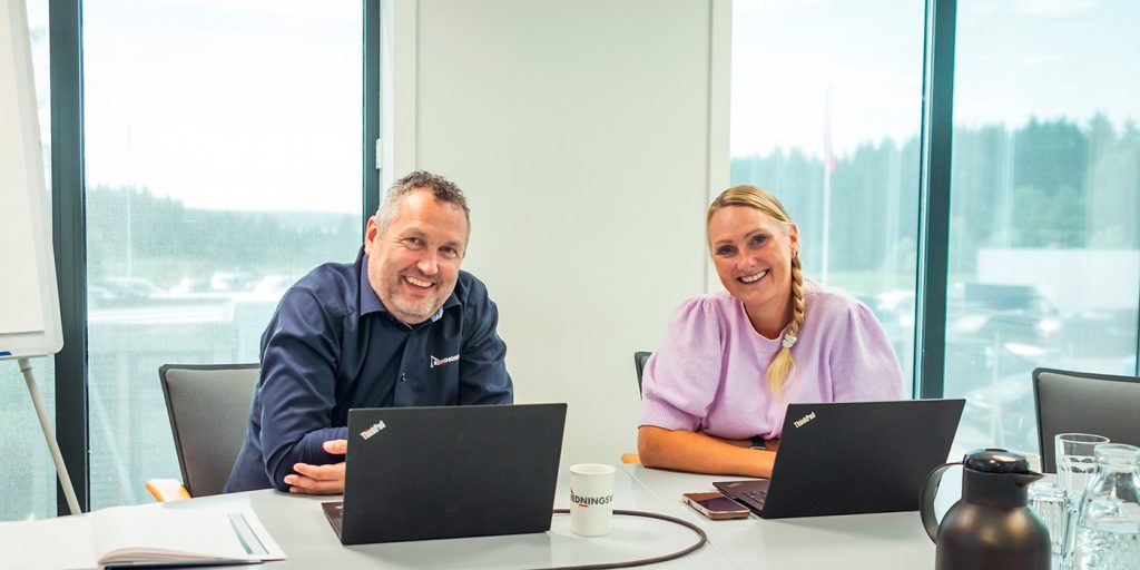 To personer sitter ved et konferansebord med bærbare datamaskiner og smiler til kameraet. På bordet står en kaffekopp, en termos og Redningsverkets materiell, og store vinduer danner en lys og innbydende bakgrunn.