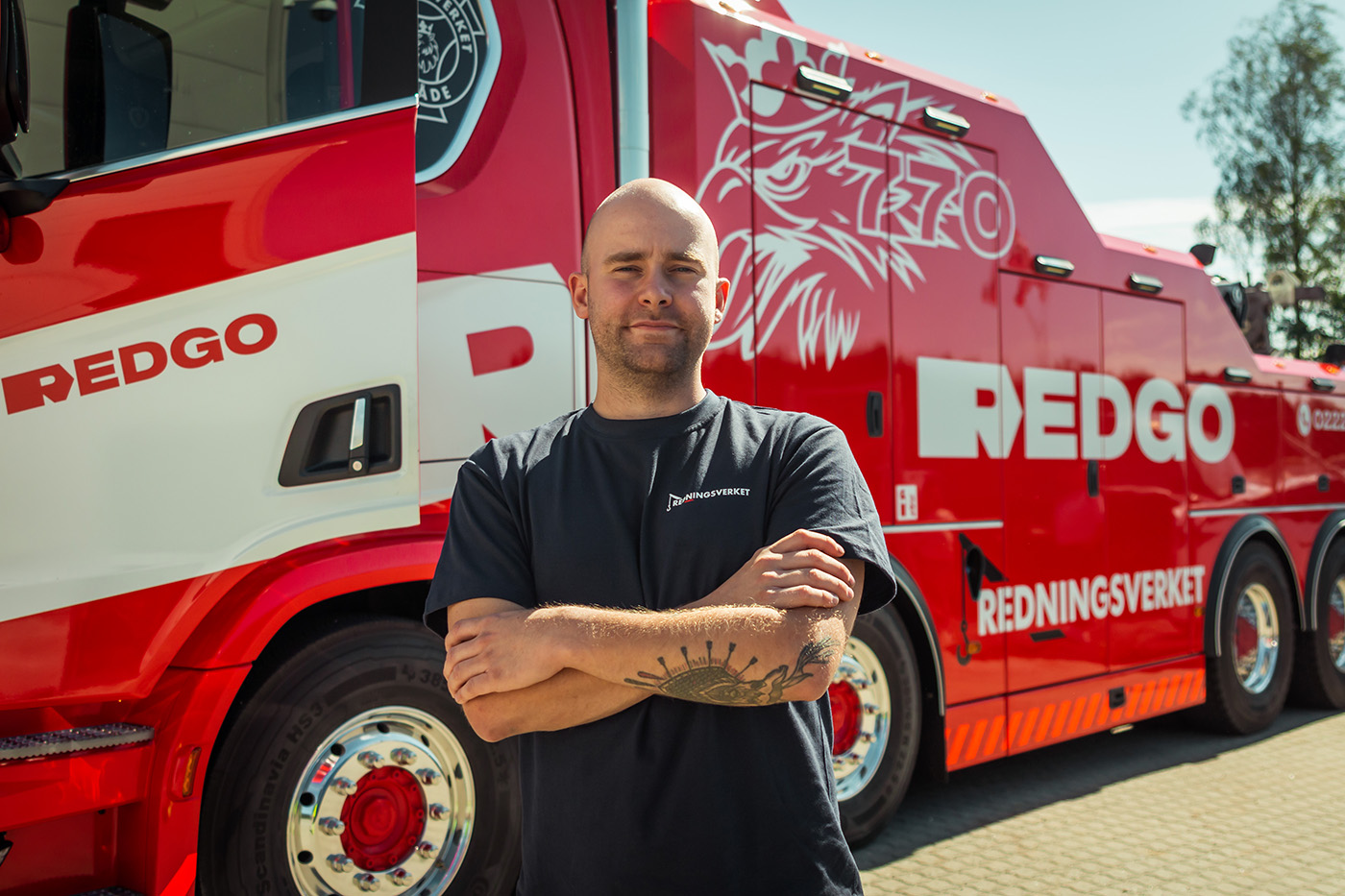 En person står med armene i kors foran en stor, rød bergingsbil med "Redningsverket" skrevet på siden.
