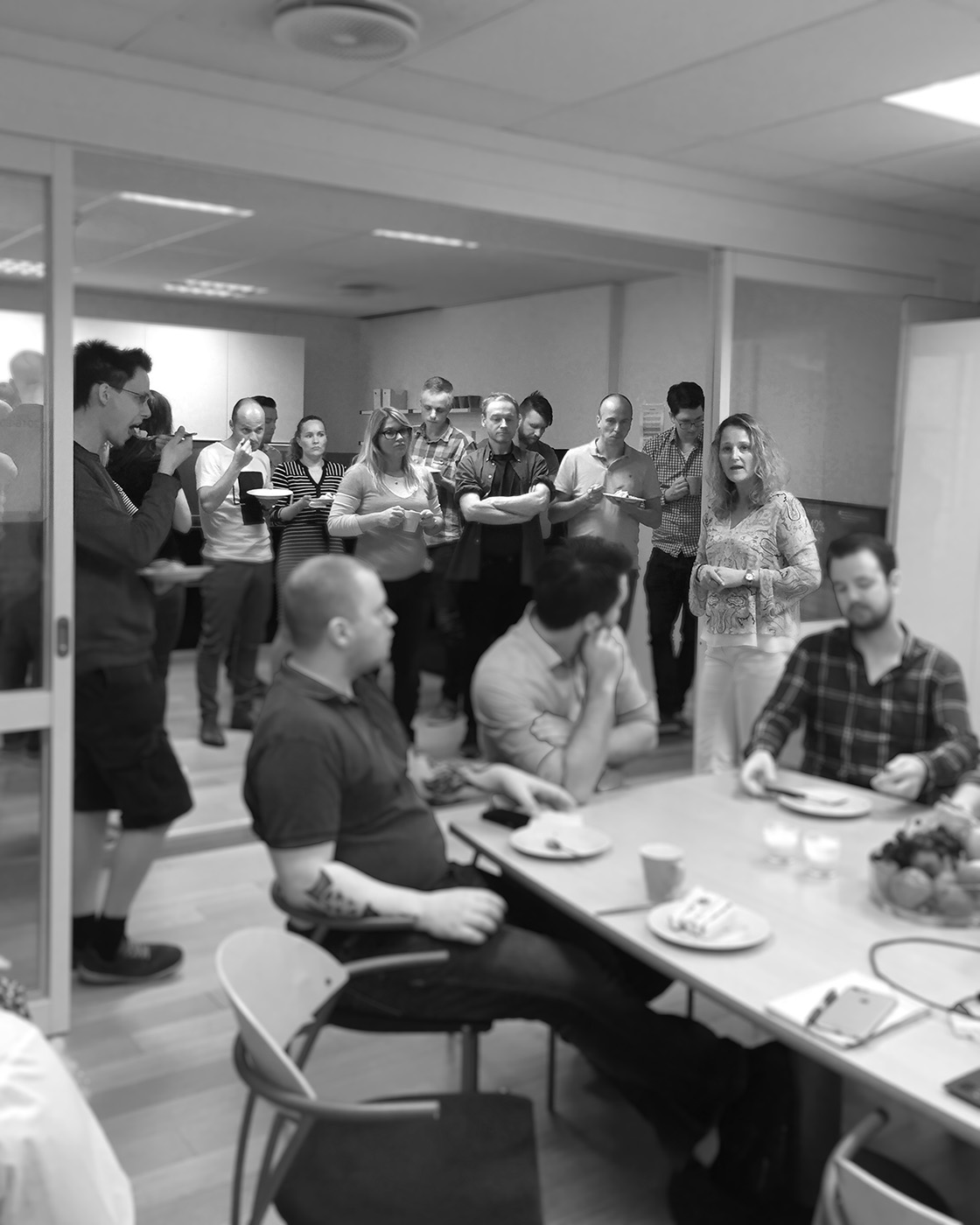 En gruppe mennesker samles i et kontorlokale, noen sitter ved et bord og spiser, mens andre står i bakgrunnen og ser ut til å lytte eller snakke - for å feire at Tripletex fikk 10 000 kunder .