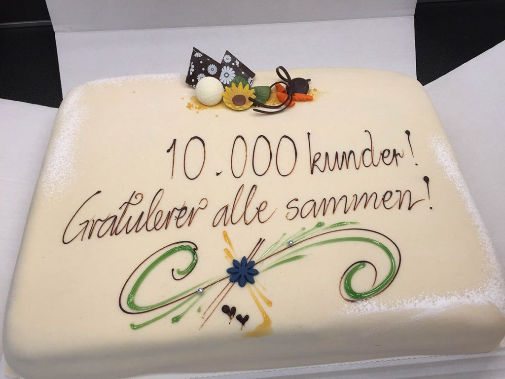 Rektangulær kake med hvit glasur, dekorert med sjokoladebiter og blomster. Tekst på norsk: "10 000 kunder! Gratulerer alle sammen!".