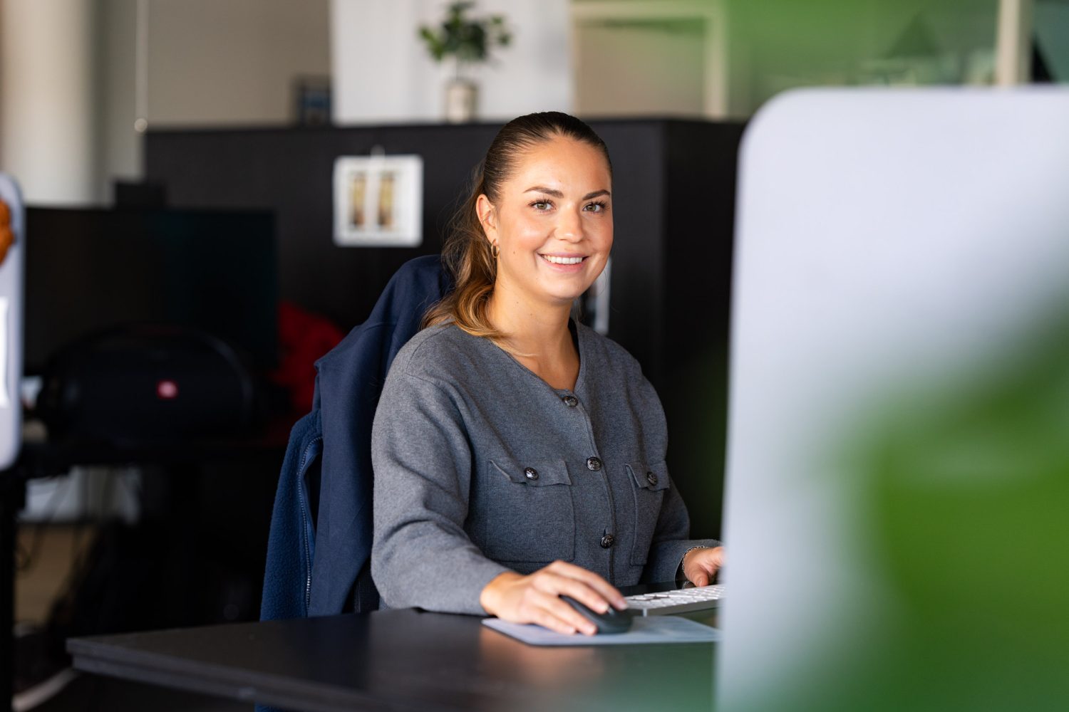 En kvinne sitter ved et skrivebord og smiler til kameraet mens hun bruker en datamaskin i et moderne kontormiljø.