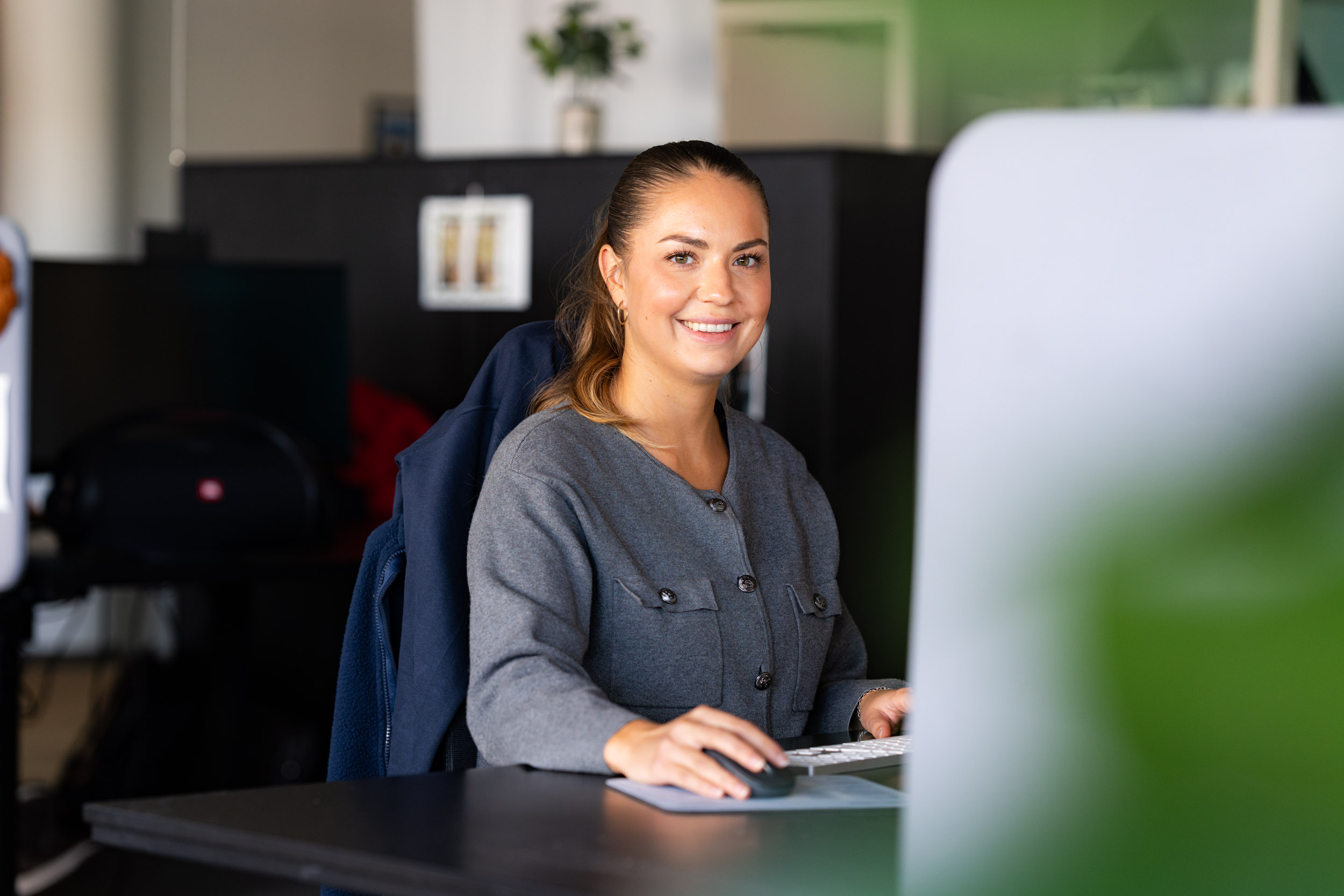 En kvinne sitter ved et skrivebord og smiler til kameraet mens hun bruker en datamaskin i et moderne kontormiljø.