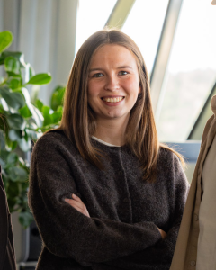 En kvinne med rett, brunt hår, iført mørk genser, står innendørs med armene i kors og smiler til kameraet.