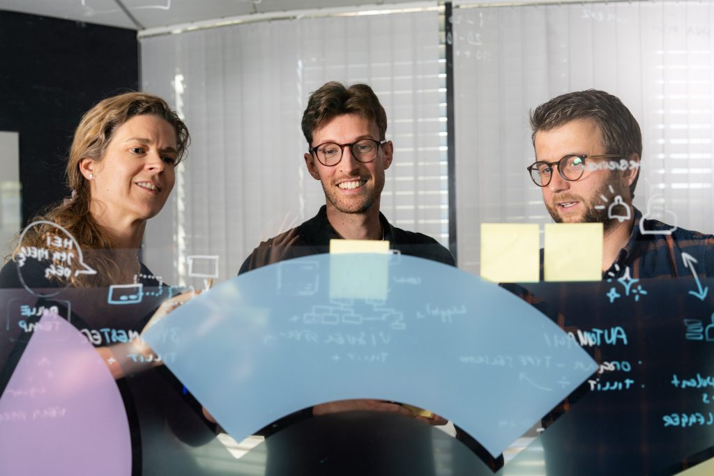 Tre personer står bak en glasstavle dekket av diagrammer og notater og diskuterer i et moderne kontormiljø.