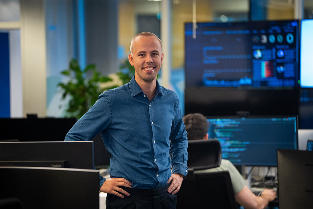 Product Manager for AI-teamet i Tripletex, Christian Hammer, er kledd i blå skjorte og står og smiler på et kontor med dataskjermer og andre mennesker som arbeider i bakgrunnen.