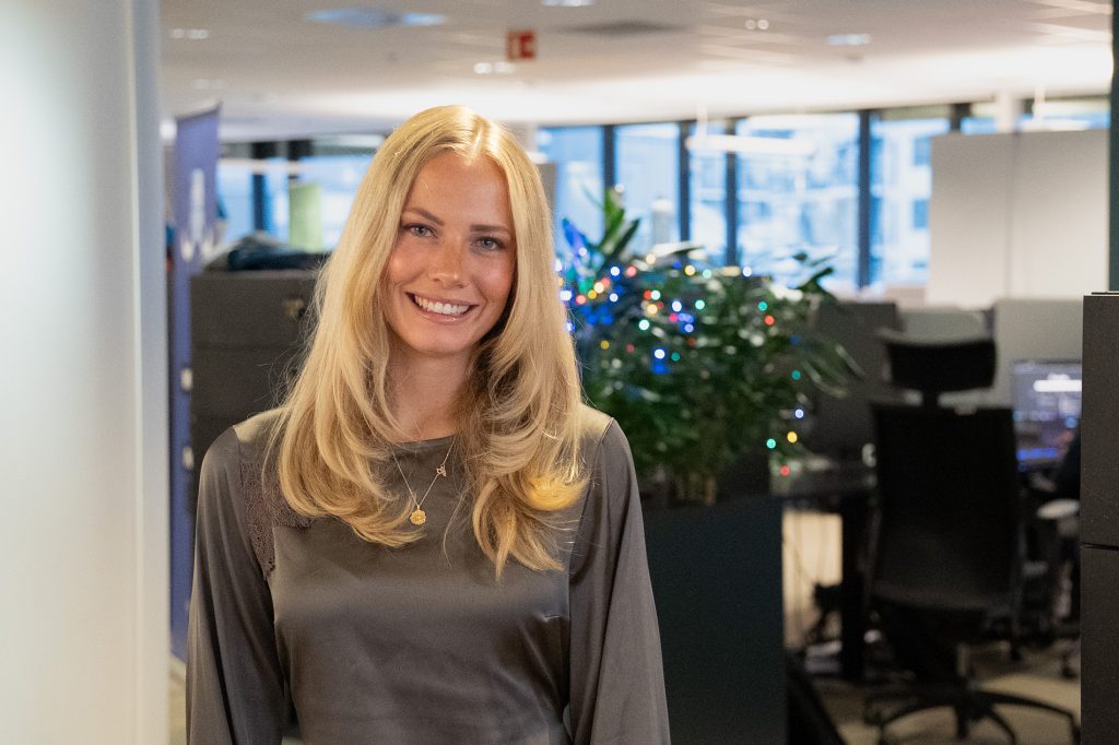 En kvinne med langt blondt hår og grå bluse står smilende på et moderne kontor med planter, dataarbeidsplasser og gaver i bakgrunnen.