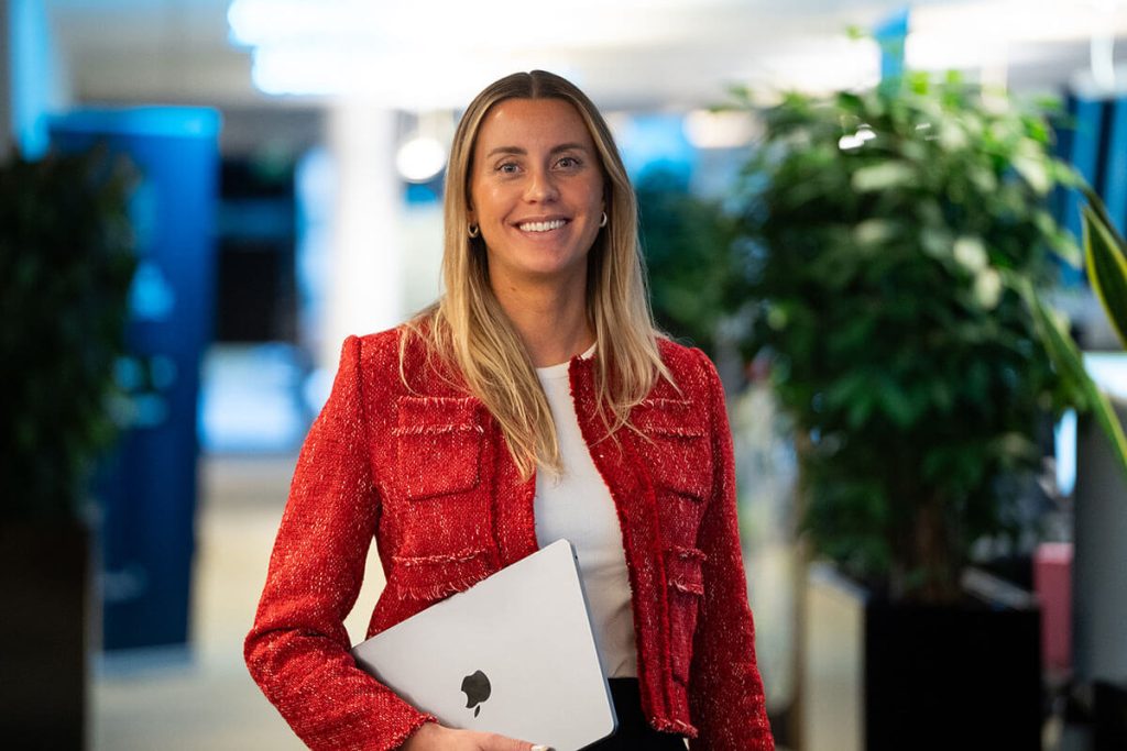 Emilie Kjønnerød, kledd i rød jakke, holder en lukket laptop mens hun står innendørs i nærheten av grønne planter og kontorbelysning.