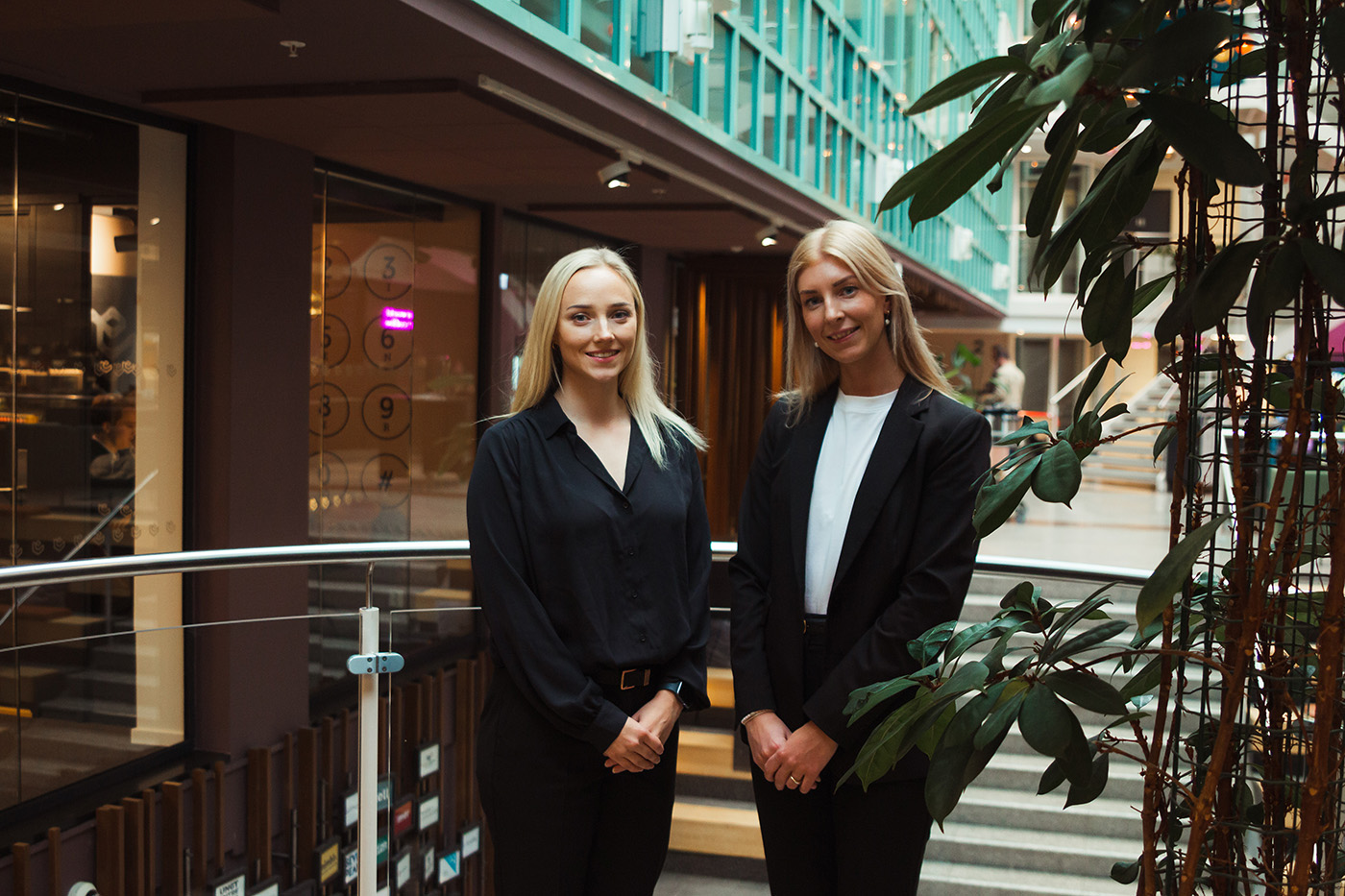 To kvinner i forretningsantrekk står innendørs ved siden av planter, med et moderne kontorbygginteriør i bakgrunnen.