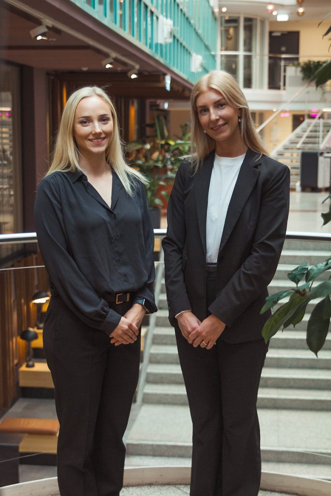 To kvinner i forretningsantrekk står side om side i et innendørs, moderne kontormiljø ved en trapp og planter.