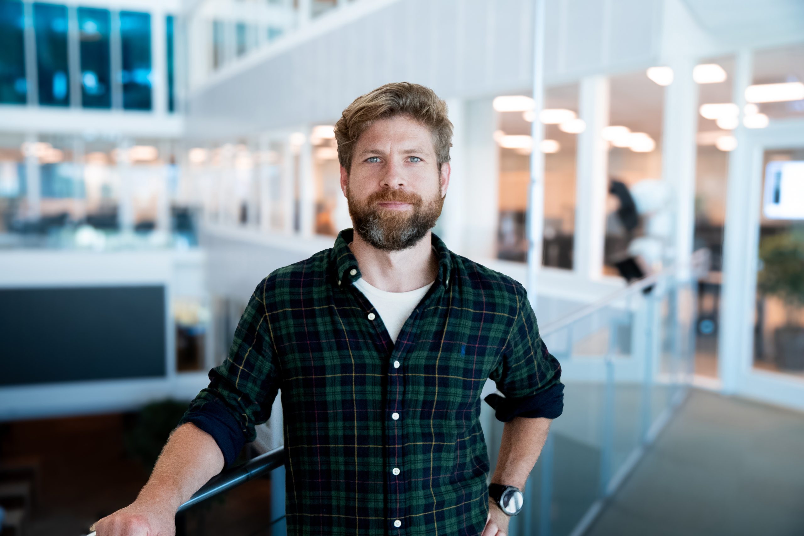En mann med skjegg og grønn rutete skjorte står i en moderne kontorbygning, hviler armen på et rekkverk og ser inn i kameraet.