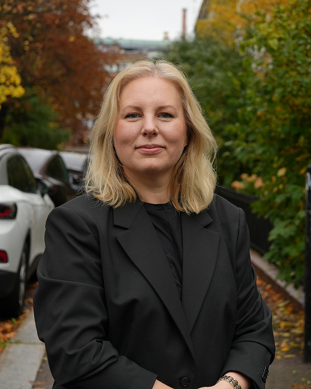 En kvinne med blondt hår og svart blazer står utendørs på et fortau, med parkerte biler og grønt løvverk i bakgrunnen.