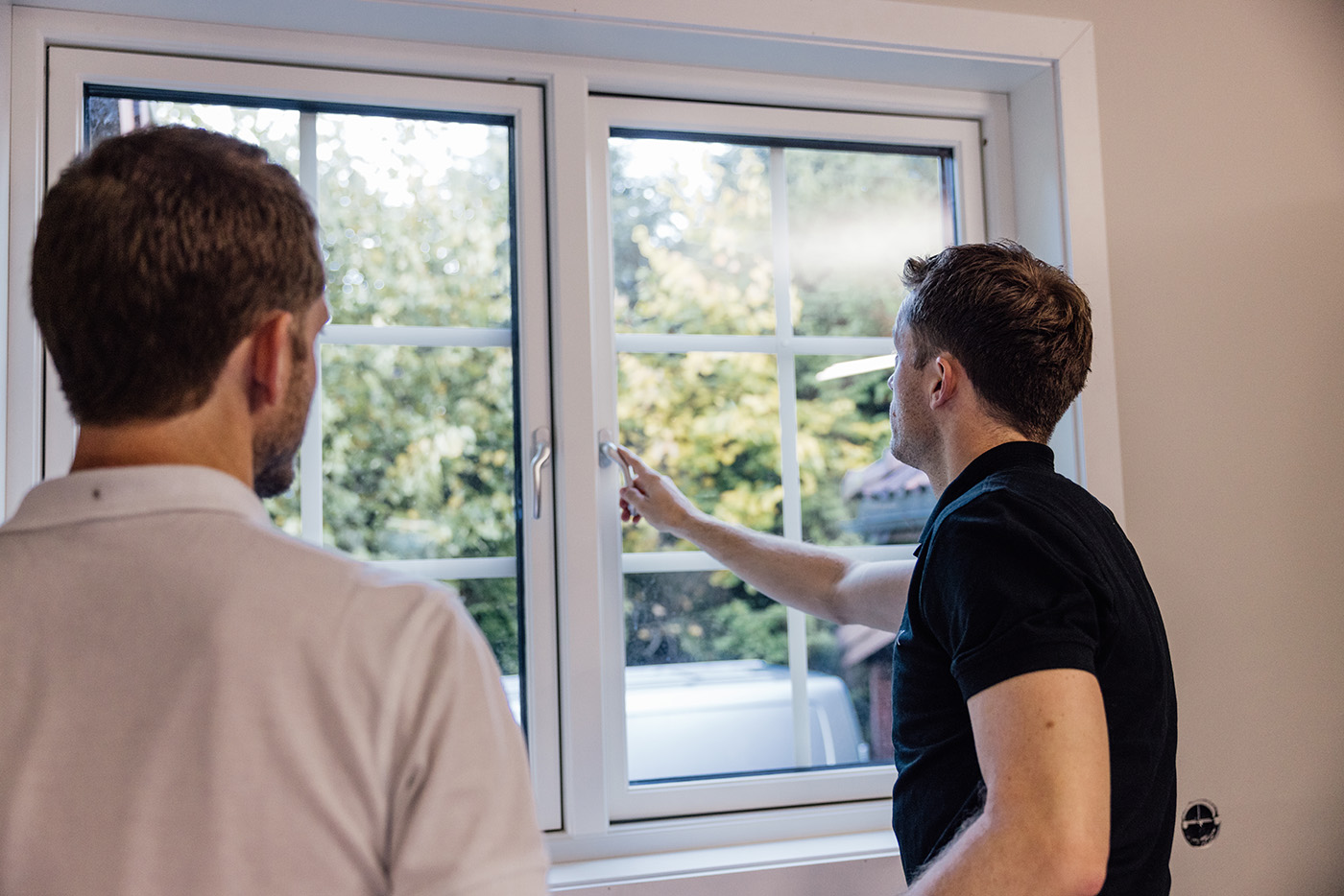 To menn står innendørs ved et lukket vindu; den ene peker ut, mens den andre ser i samme retning. Trær og et kjøretøy er synlige gjennom glasset.