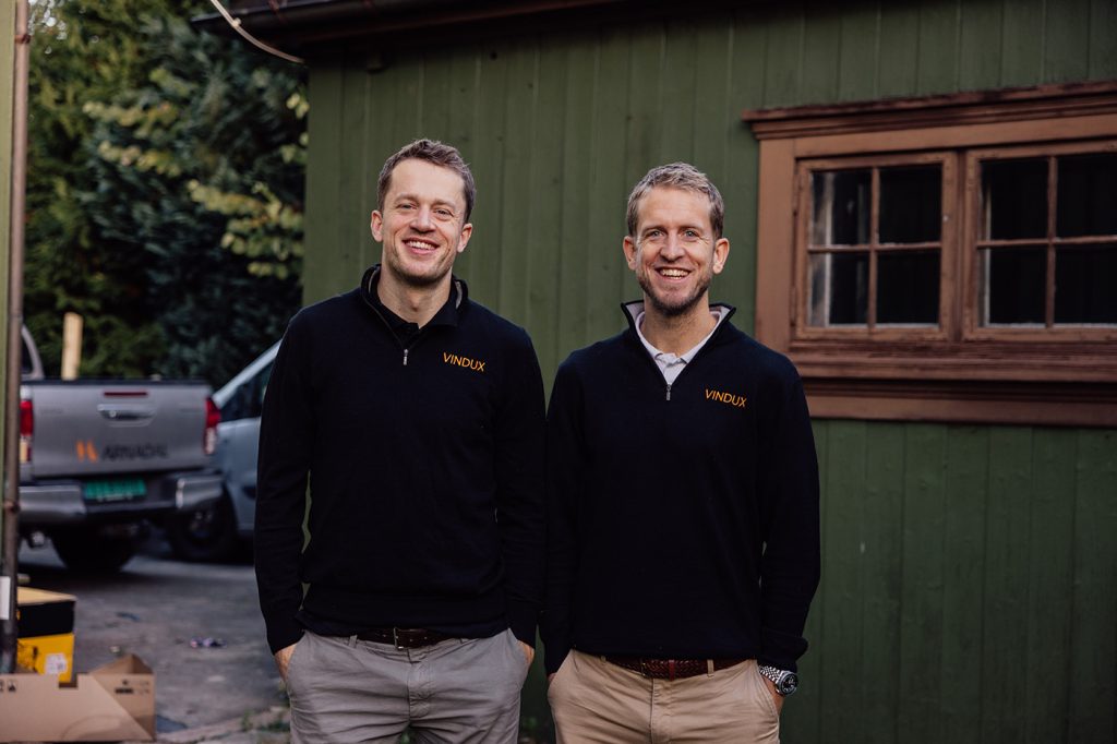 To menn iført matchende svarte VINDUX-gensere står smilende side om side foran en grønn trebygning med en lastebil parkert i nærheten. Tarjej Bø og Rasmus Endre Bø driver Vindux og bruker Tripletex.