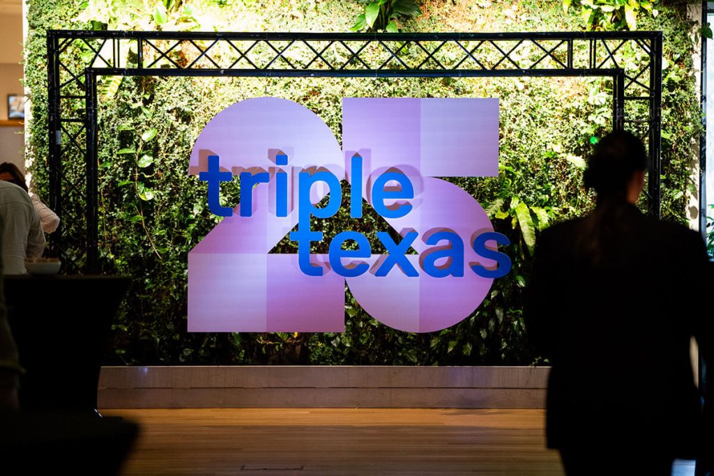 Stort skilt med teksten "Tripletexas 25" står mot en grønn plantevegg, med mennesker synlige i forgrunnen.