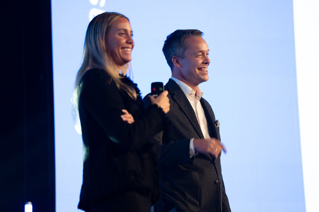 En kvinne og en mann står på scenen, smilende og med en mikrofon i hånden, foran en stor blå og hvit skjerm.
