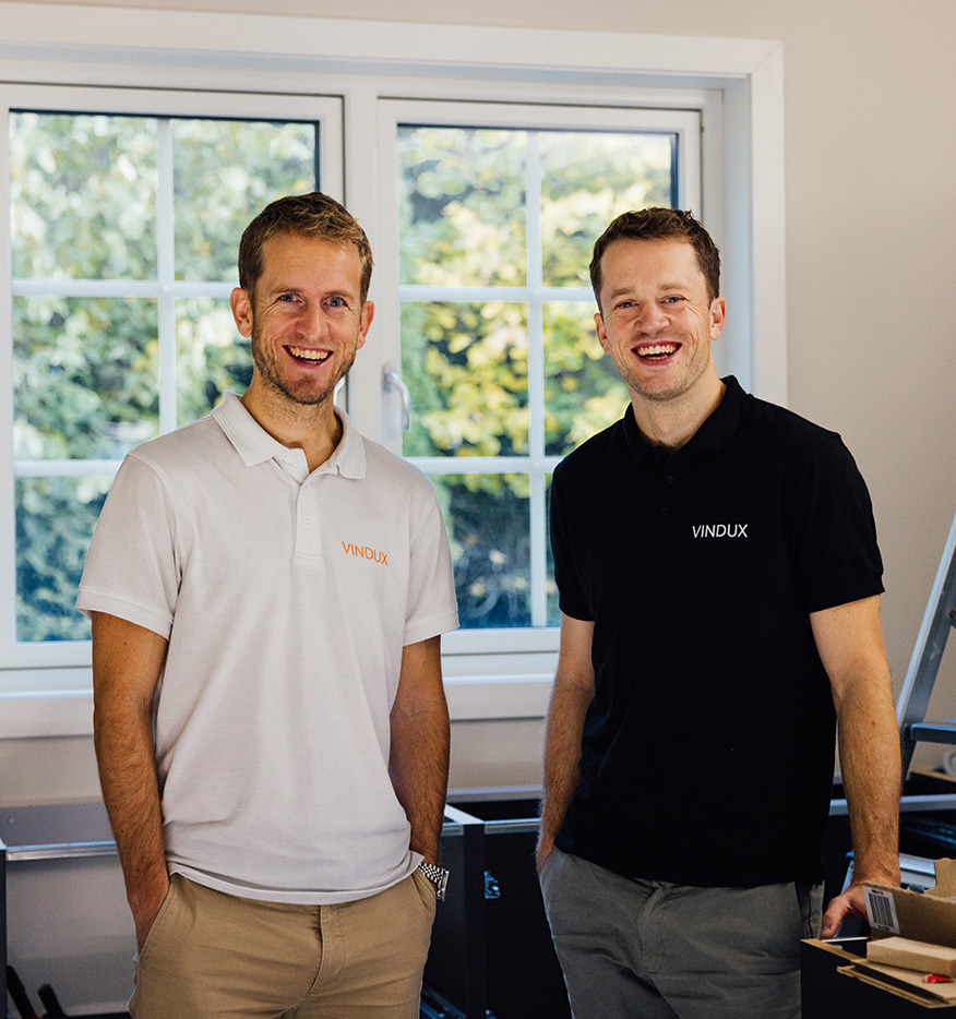 To menn iført poloskjorter med "VINDUX"-logo står og smiler i et lyst rom med et vindu og utendørs grøntområder i bakgrunnen.