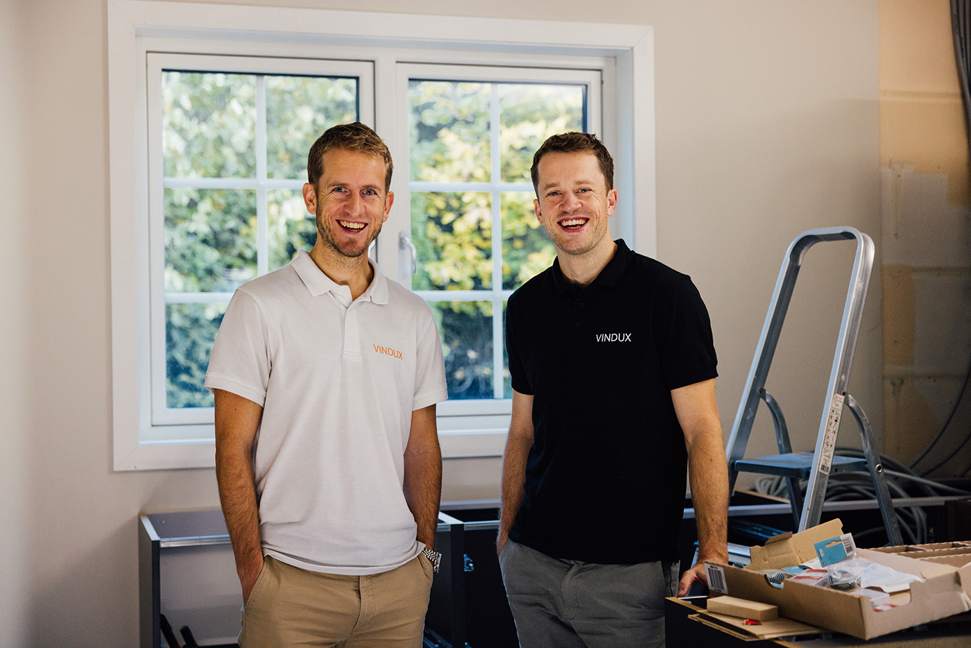 To menn står innendørs foran et vindu og smiler. De har på seg poloskjorter med merkevare og er omgitt av esker og utstyr.