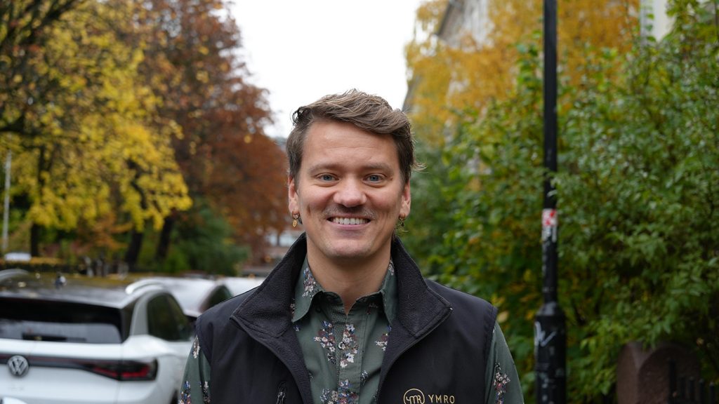 En person med kort hår og blomstrete skjorte står ute i en gate med høsttrær og parkerte biler og smiler til kameraet.