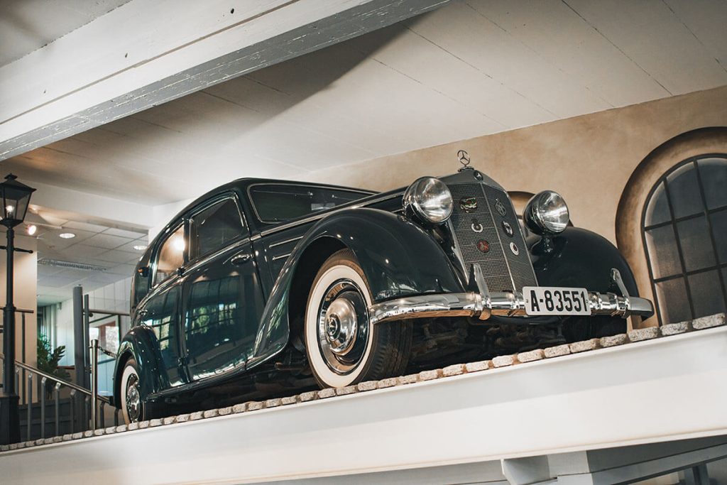 En svart luksusbil med hvite dekk står utstilt på en forhøyet plattform innendørs, under kunstig belysning.