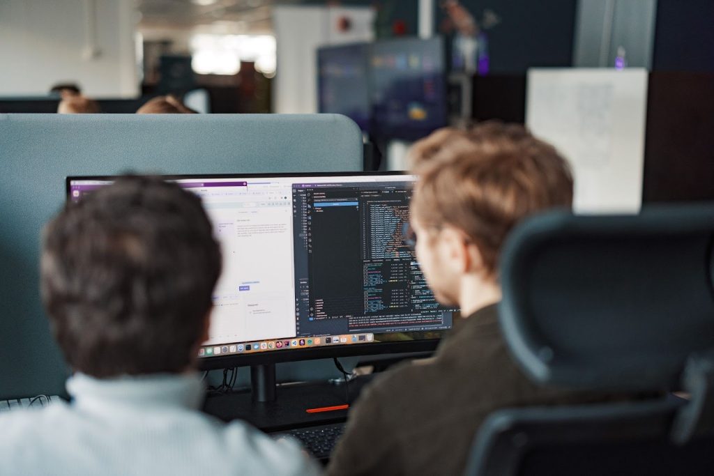To personer sitter ved et skrivebord og ser på dataskjermer som viser kode og nettsider i et kontormiljø.