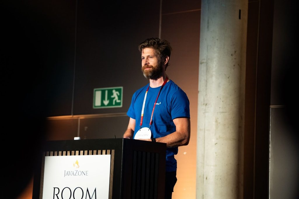 En mann i blå t-skjorte står på et podium merket "JavaZone ROOM" og holder en presentasjon i en konferansesetting.
