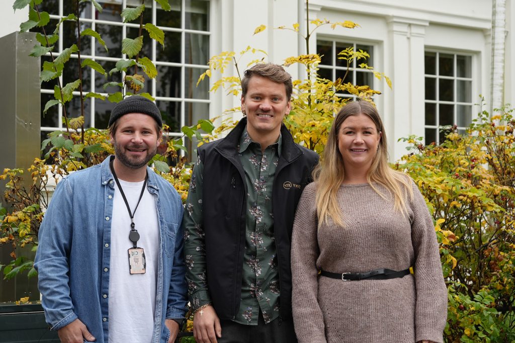 Tre personer står utendørs foran en hvit bygning med store vinduer, omgitt av gulgrønt løvverk.