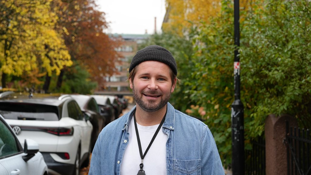 En mann iført svart lue og denimskjorte står utendørs i en gate med parkerte biler og høsttrær i bakgrunnen.