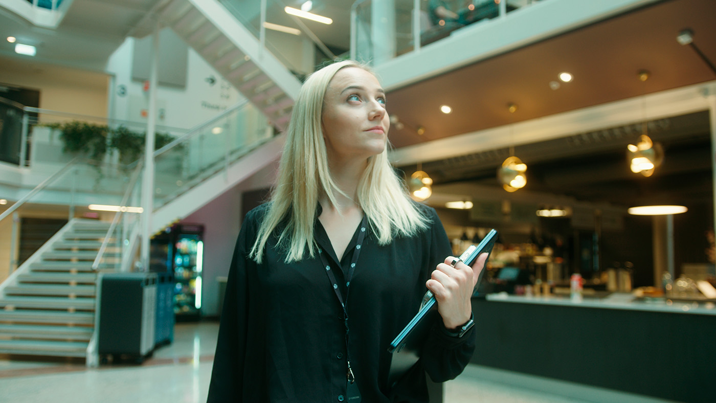 En kvinne med langt blondt hår holder et nettbrett og ser opp mens hun står i en moderne kontorlobby med trapper og moderne belysning. Cathrine Ro Heuch driver Nordic Brain Tech.