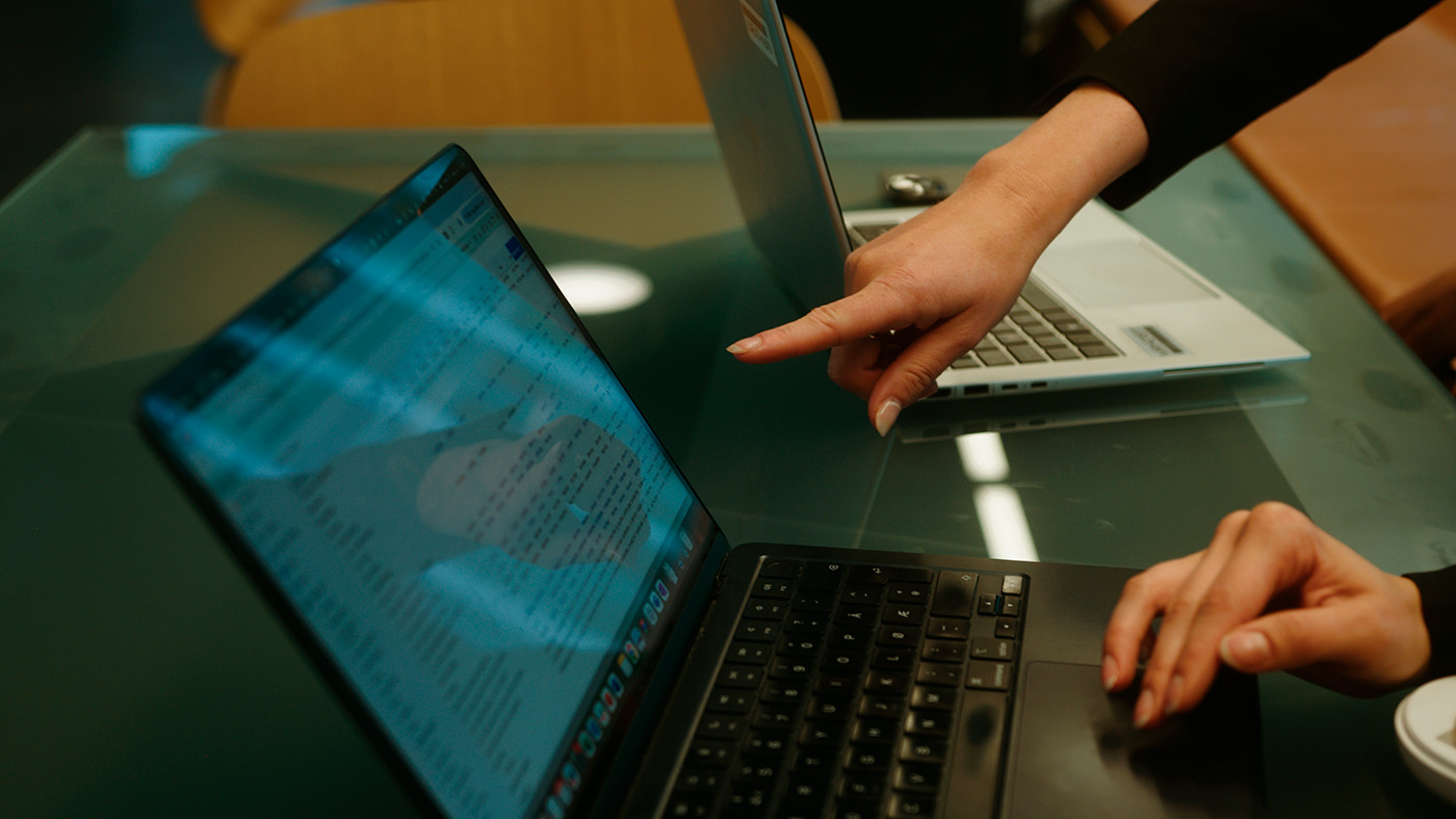 To kvinner i forretningsantrekk står innendørs ved siden av planter, med et moderne kontorbygginteriør i bakgrunnen.