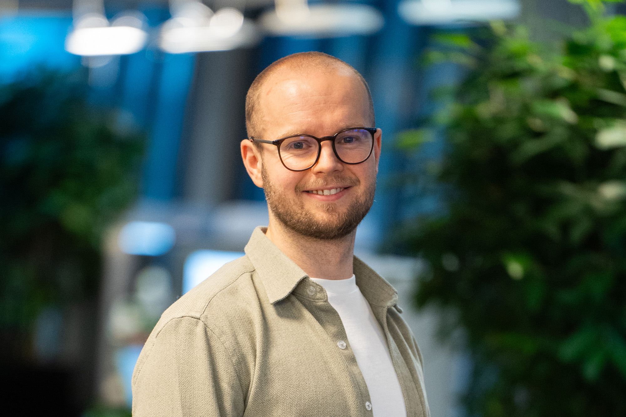 En mann med briller og skjegg smiler mens han står innendørs, iført en lys skjorte over en hvit t-skjorte. I bakgrunnen er det grønne planter.