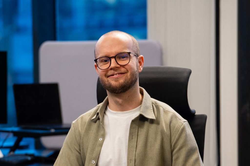 En mann med briller og skjegg, iført grønn skjorte over en hvit t-skjorte, sitter og smiler i et kontormiljø med datamaskiner i bakgrunnen. Thomas Lyseggen er Tech lead i Tripletex.