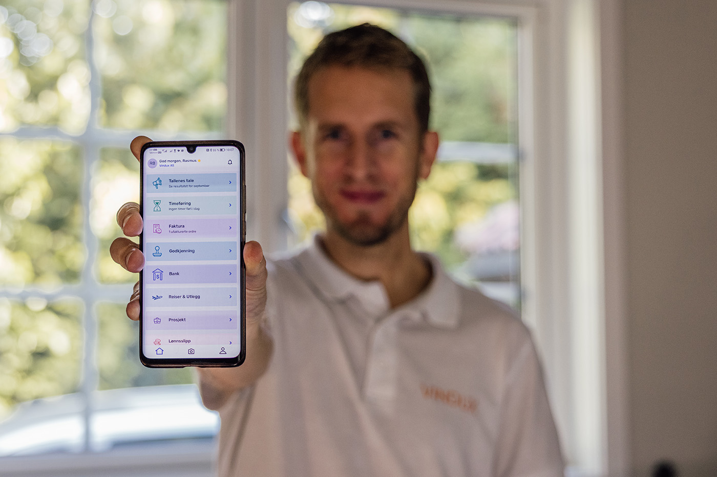 En mann i hvit poloskjorte holder en smarttelefon mot kameraet og viser et grensesnitt for en mobilapp; ansiktet hans er ute av fokus.